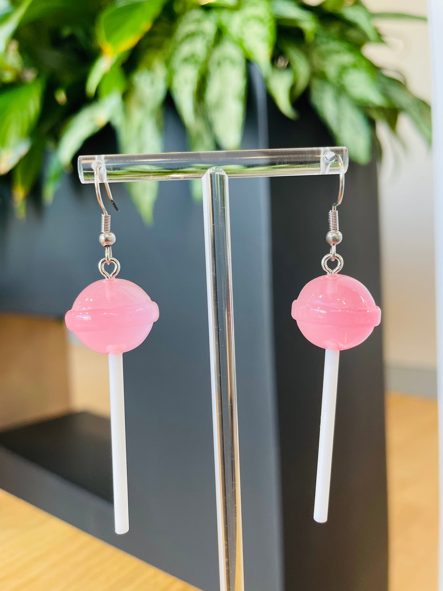 A pair of lollipop-shaped earrings from Playful Pantry in pink color displayed on an earing stand, with a green plant in the background.