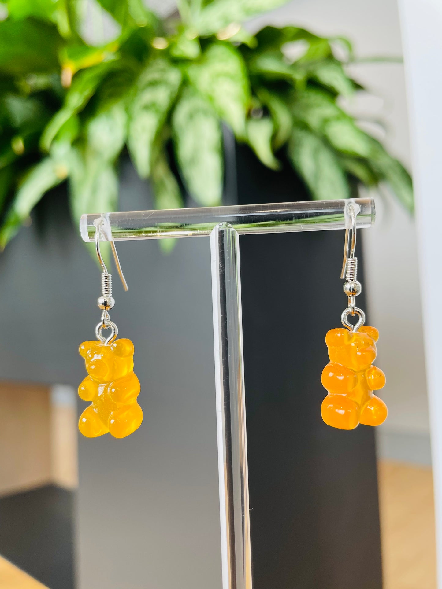 A pair of yellow candy-shaped earrings displayed on an ear stand, with a blurred background featuring green foliage.