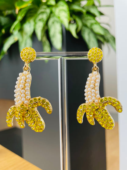 A pair of banana-shaped earrings adorned with yellow and white gems on a metal structure, displayed against a green leafy background.