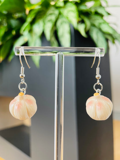 A pair of garlic-shaped earrings displayed on a clear stand with a green plant in the background.