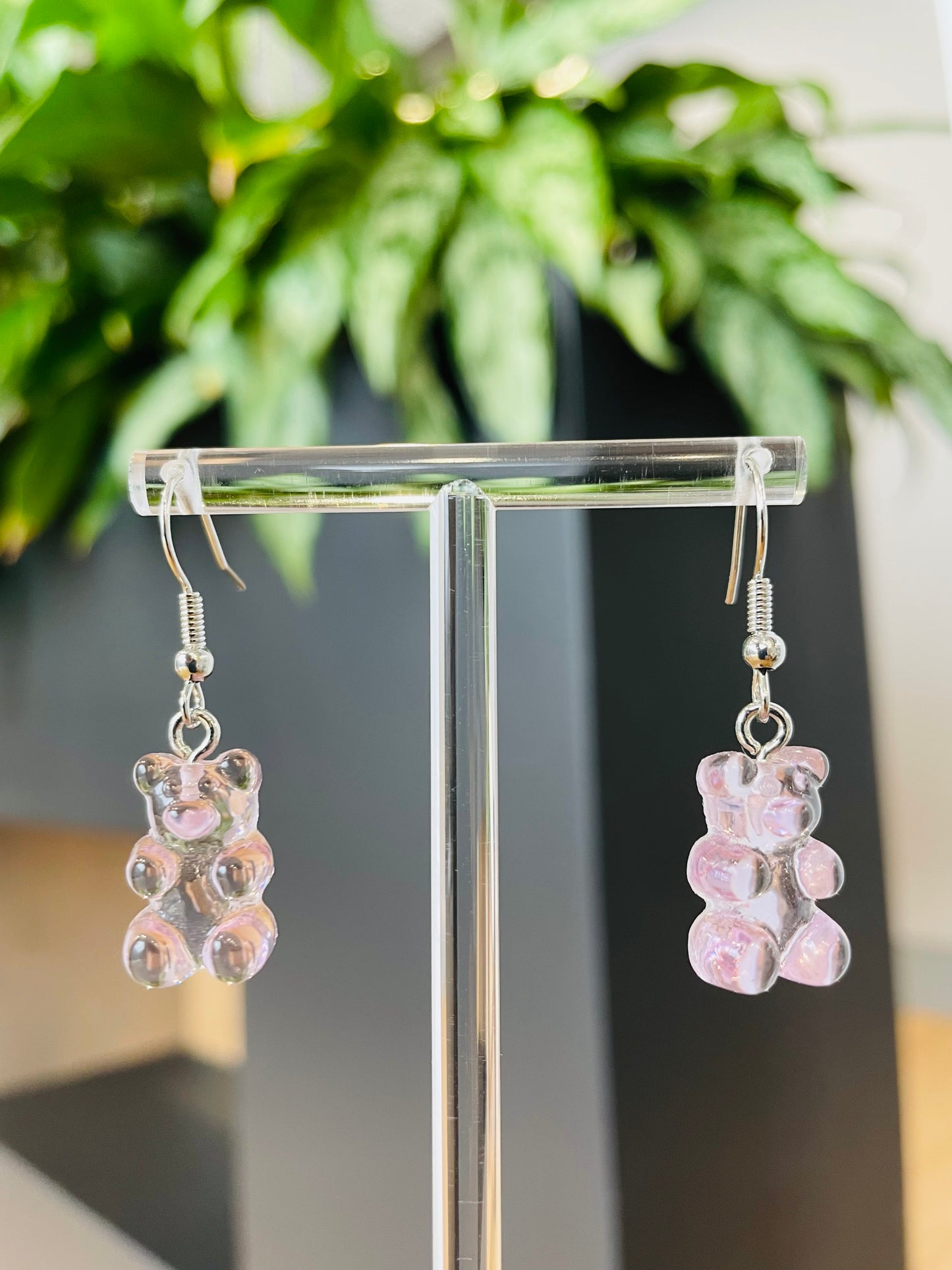 A pair of lilac candy-shaped earrings displayed on an ear stand, with a blurred background featuring green foliage.
