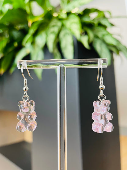 A pair of lilac candy-shaped earrings displayed on an ear stand, with a blurred background featuring green foliage.