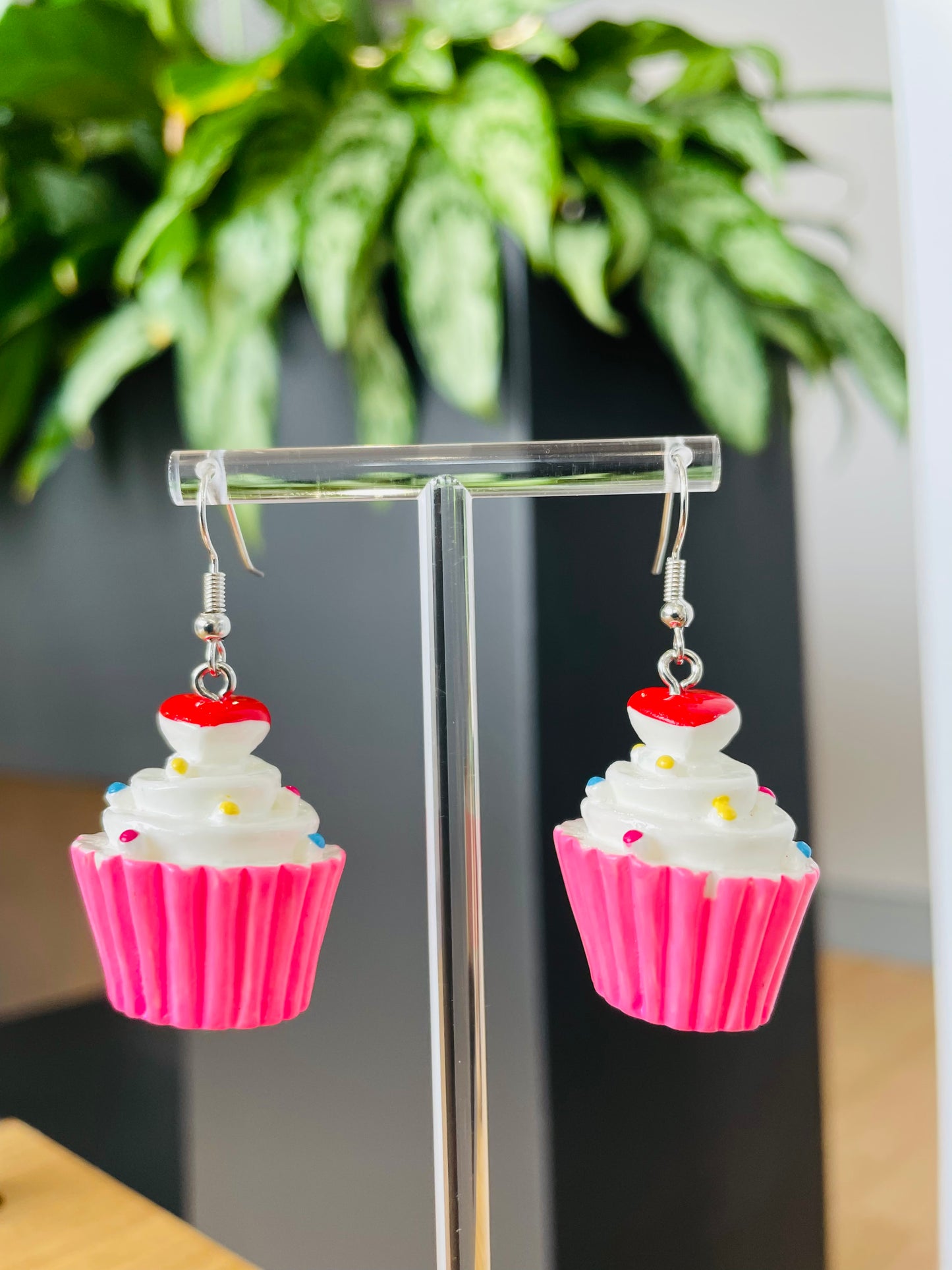 A pair of pink cupcake-shaped earrings with white frosting and red sprinkles displayed on a stand, with a green plant in the background.