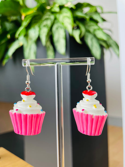 A pair of pink cupcake-shaped earrings with white frosting and red sprinkles displayed on a stand, with a green plant in the background.