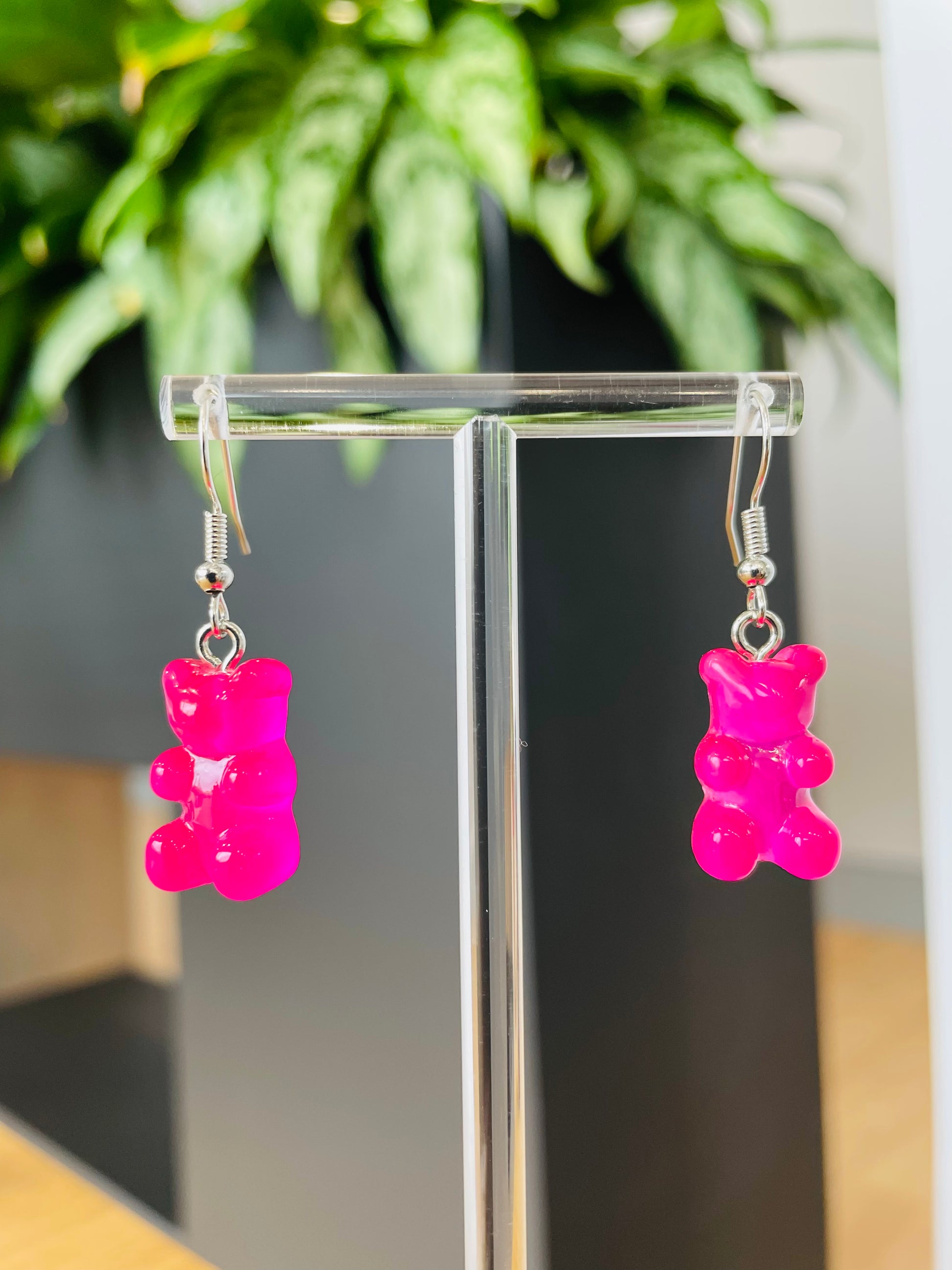 A pair of pink candy-shaped earrings displayed on an ear stand, with a blurred background featuring green foliage.