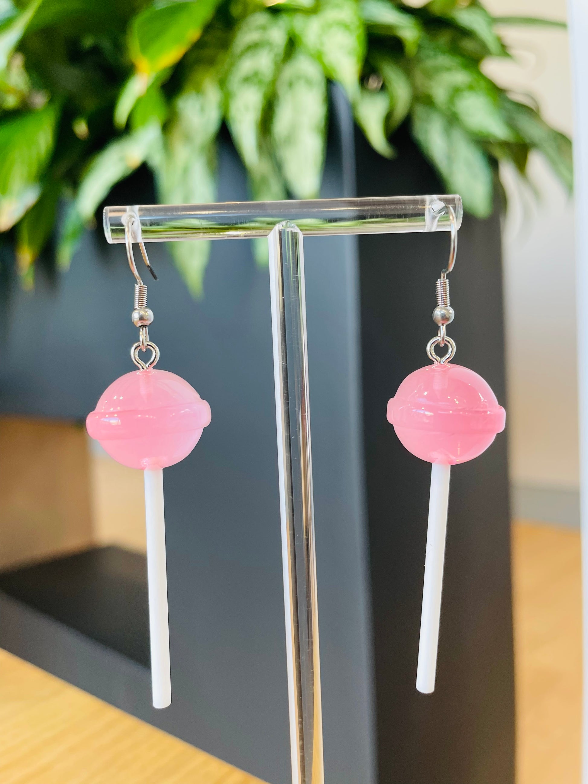 A pair of lollipop-shaped earrings from Playful Pantry in pink color displayed on an earing stand, with a green plant in the background.