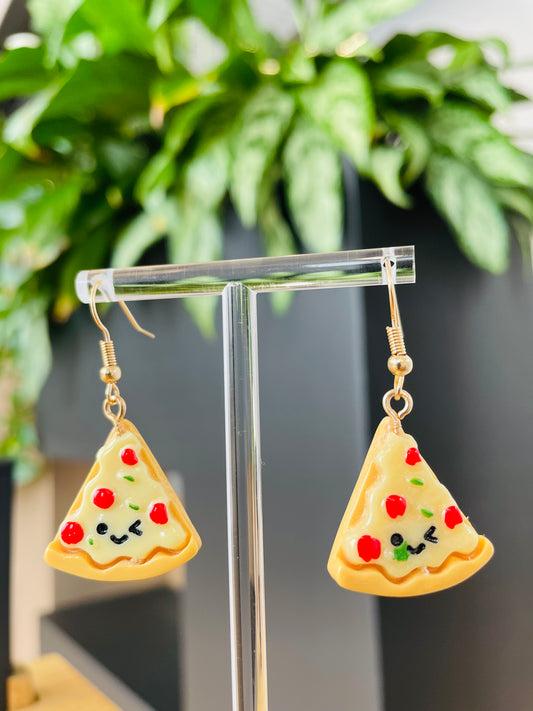 A pair of pizza slice earrings displayed on a stand, featuring yellow and red colors with black accents, resembling a cheese pizza with toppings.