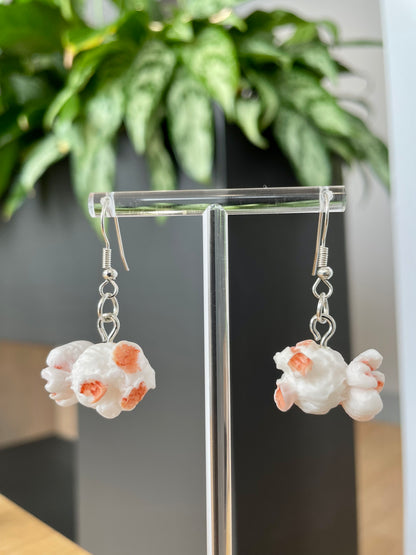 A pair of sweet popcorn-shaped earrings displayed on an acrylic stand with a green plant in the background.