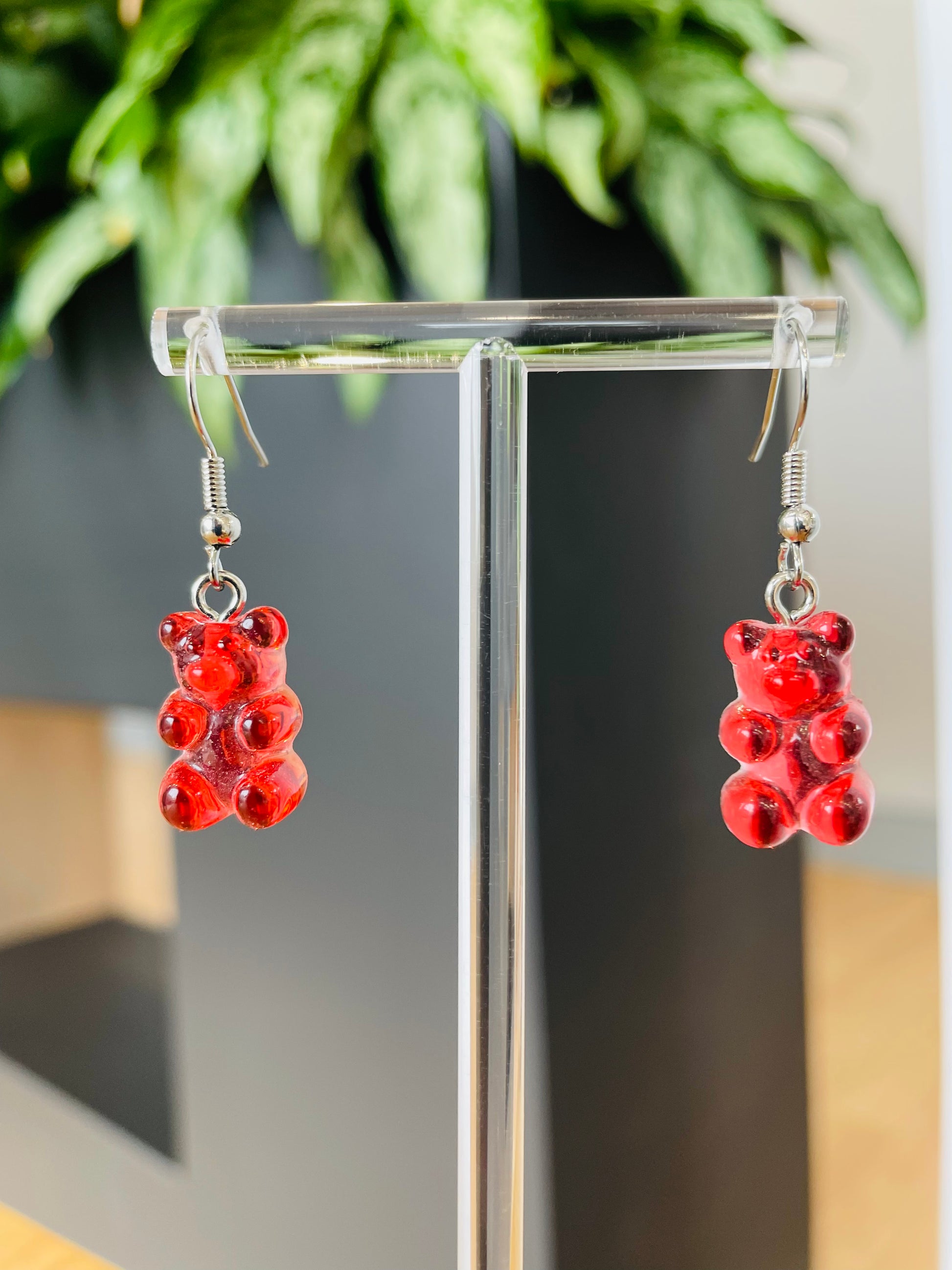 A pair of red candy-shaped earrings displayed on an ear stand, with a blurred background featuring green foliage.