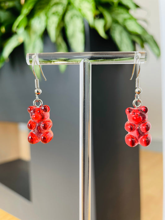A pair of red candy-shaped earrings displayed on an ear stand, with a blurred background featuring green foliage.