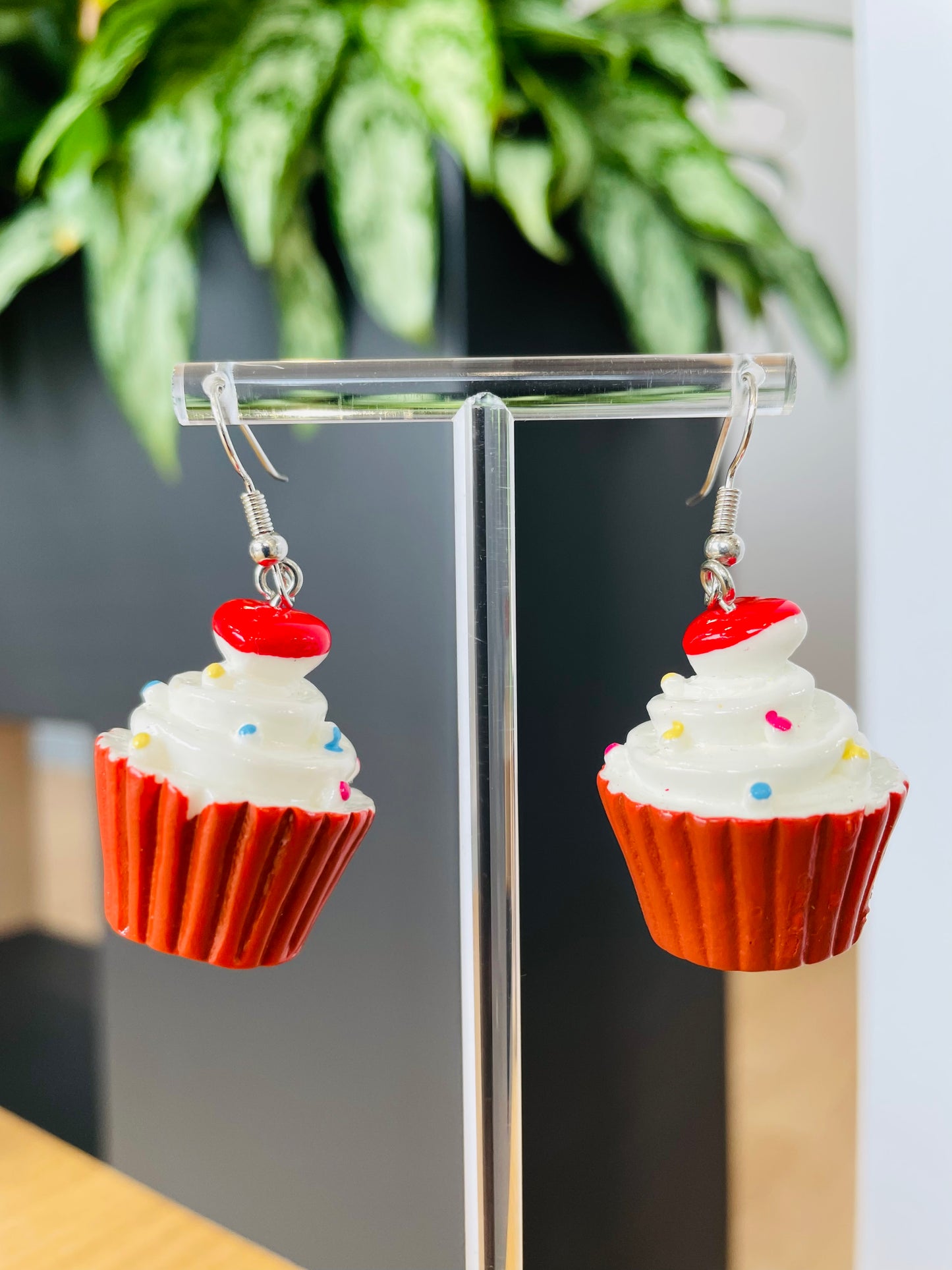 A pair of red cupcake-shaped earrings with white frosting and red sprinkles displayed on a stand, with a green plant in the background.