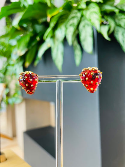 A pair of red strawberry stud earrings displayed on a stand with a plant in the background.