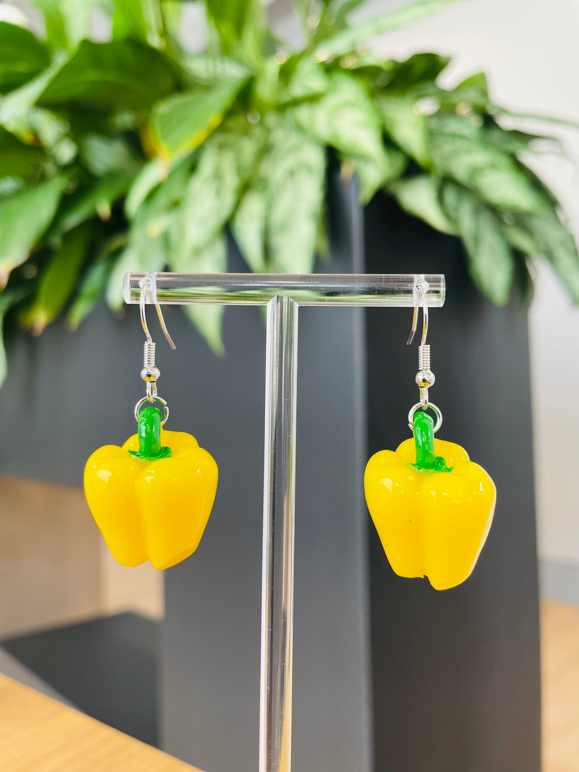 A pair of vibrant yellow bell pepper earrings displayed on an earring stand, with green accents representing the stem.