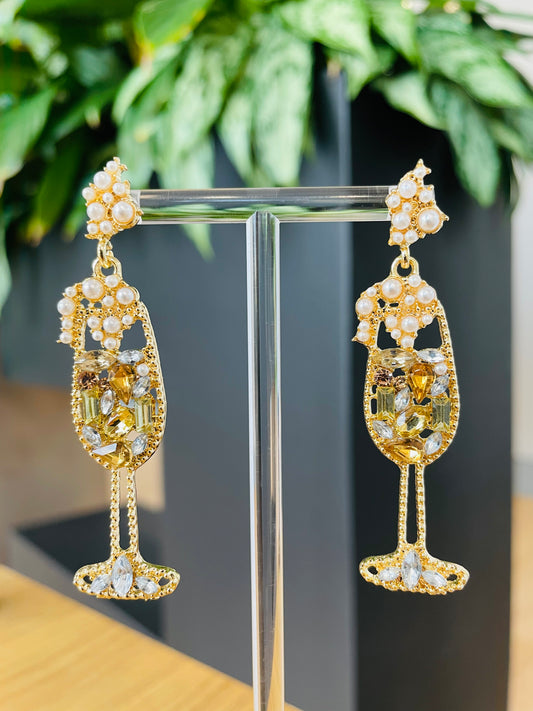 A pair of champagne glass-shaped earrings displayed on a stand, featuring gold color with embellishments.
