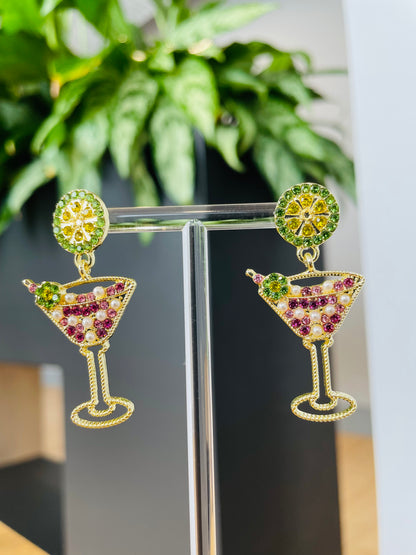 A pair of gold-colored martini glass earrings with pink and green accents displayed on a stand, with a plant in the background.