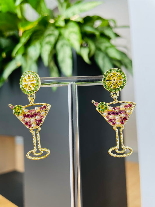 A pair of gold-colored martini glass earrings with pink and green accents displayed on a stand, with a plant in the background.